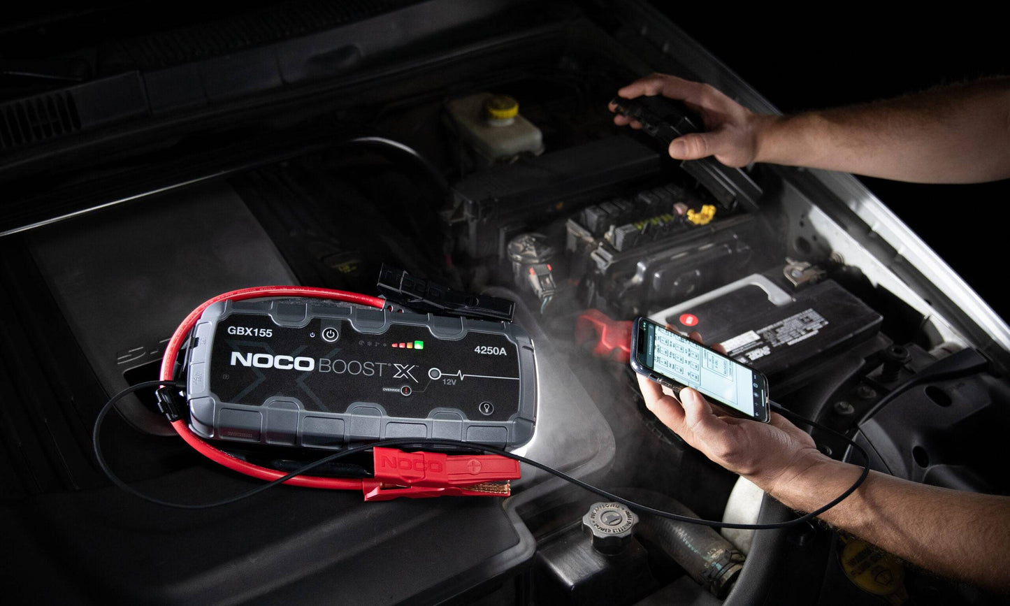 Noco Boost X GBX155 4250A jump starter in use under the bonet, jump-starting a vehicle battery while charging a phone, perfect for roadside emergencies.