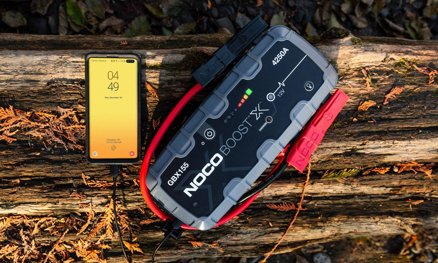 Noco Boost X GBX155 Jump Starter 4250A charging a smartphone outdoors, ideal for emergency power on the go for vehicles and mobile devices.